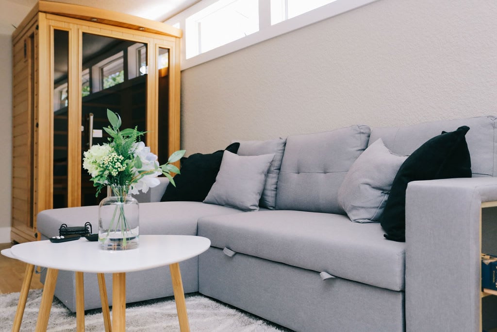 A comfortable sectional sofa in light gray is adorned with black and patterned throw pillows. A small, round coffee table sits in front, featuring a vase with fresh flowers. Large windows allow natural light to fill the welcoming living space.