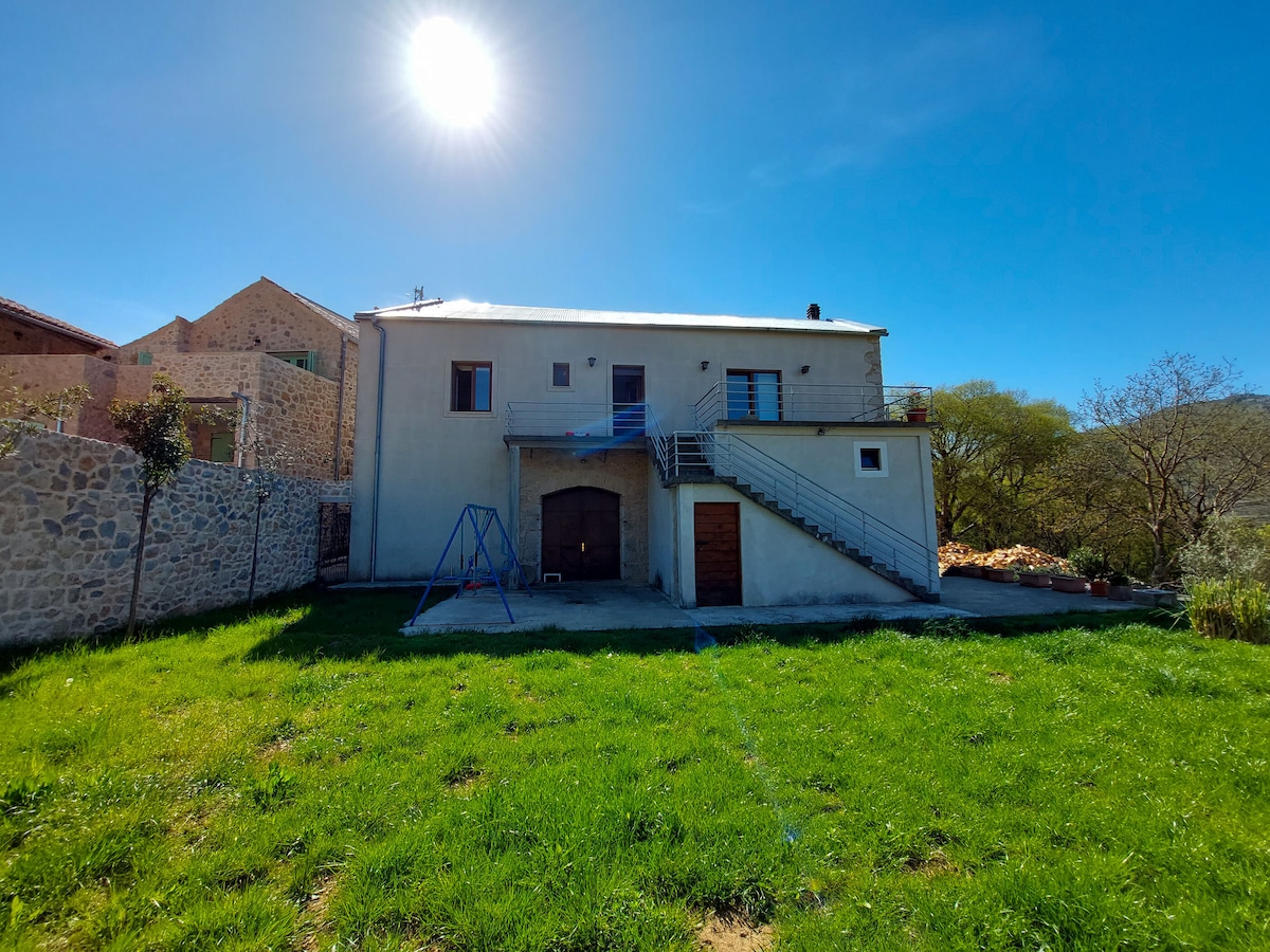 A spacious building stands under a bright sun, featuring a staircase leading to an upper level. A swing set is visible in the grassy yard, alongside stone walls and trees in the background, creating a sense of open space and tranquility.