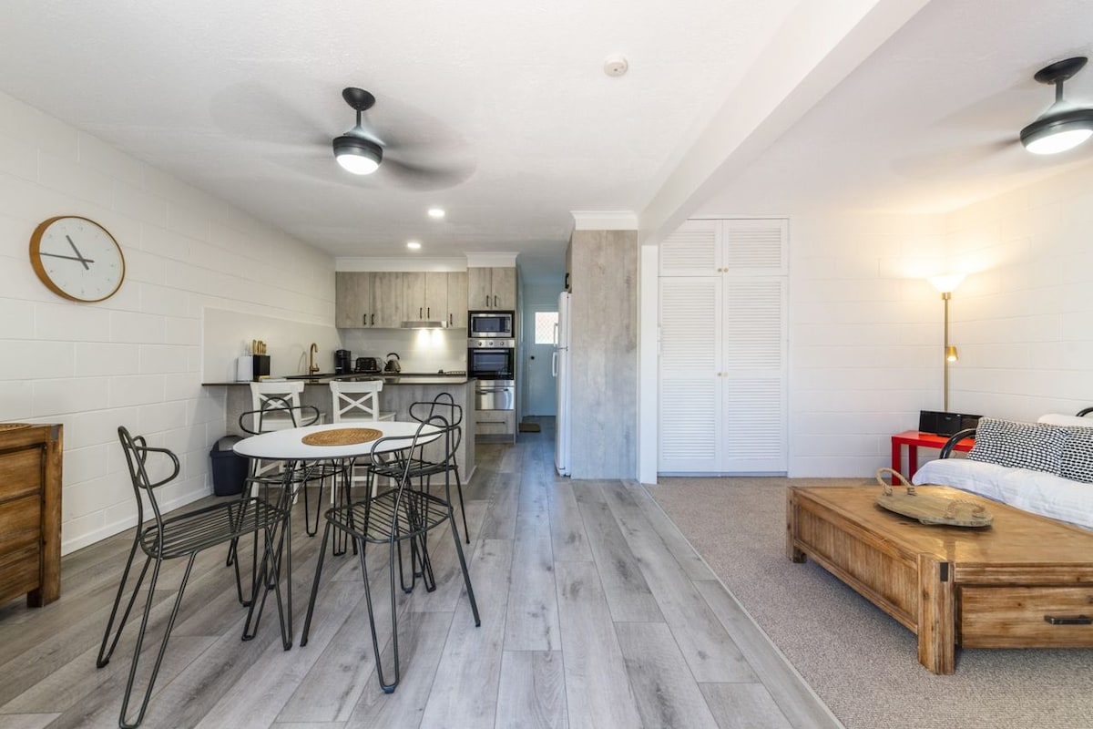 A modern living space is presented, featuring a stylish dining area with a round table and six chairs. The open kitchen showcases sleek cabinets and new appliances. Natural light fills the room, highlighted by a ceiling fan and a spacious seating area with wooden accents.