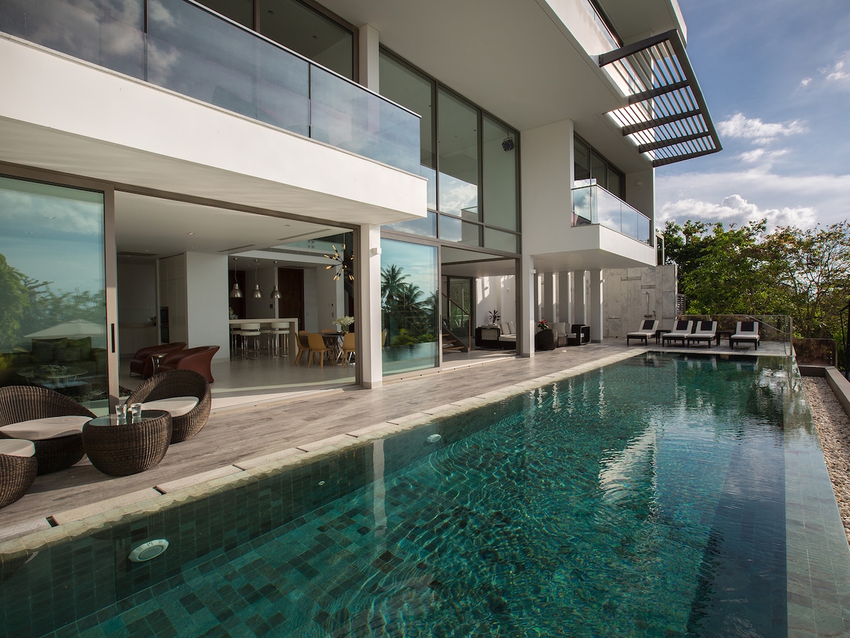 The outdoor area features a sleek infinity pool with crystal-clear waters, surrounded by modern lounge chairs. Glass doors provide views into the open-plan living space, which is bathed in natural light from ample windows, blending seamlessly with the surrounding greenery.
