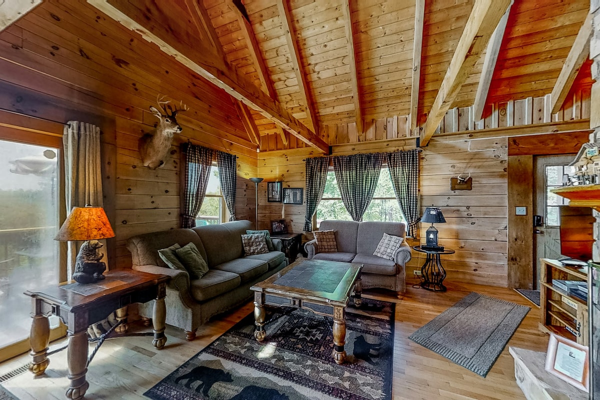 The living area features high, exposed wood-beamed ceilings and large windows that offer natural light and views. Comfortable seating is arranged around a wooden coffee table, with a patterned area rug beneath. Decorative accents include a lamp and wall art, enhancing the rustic cabin feel.