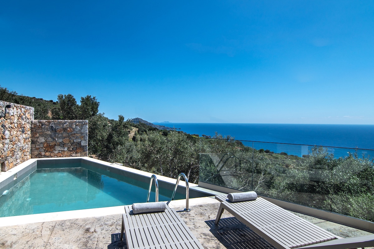 A private swimming pool is framed by stone walls, offering unobstructed views of the sea and hills. Sun loungers are positioned alongside the pool, inviting relaxation under clear blue skies.