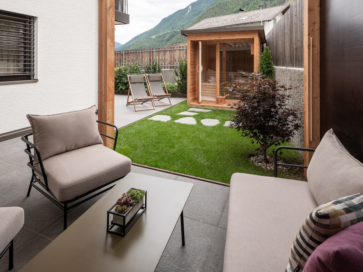 A serene outdoor space is presented, featuring a well-maintained lawn and a wooden structure that can serve as a shelter. Comfortable seating areas with soft cushions are arranged around a low table, inviting relaxation while enjoying views of the surrounding mountains.