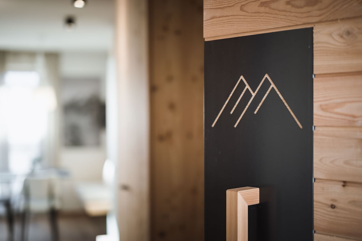 A wooden entrance area features a minimalist design with a mountain graphic etched in the wall. Soft lighting provides a warm ambiance, leading further into the living space visible in the background.
