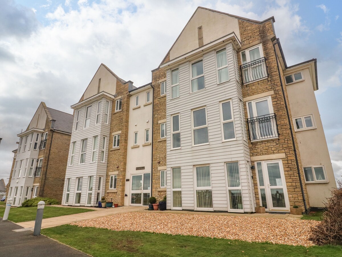 The exterior of the apartment building is showcased, featuring a combination of stone and siding. Large windows allow for natural light, while the landscaped pathway leads to the entrance. Gravel outlines the entrance area, contributing to the inviting appearance.