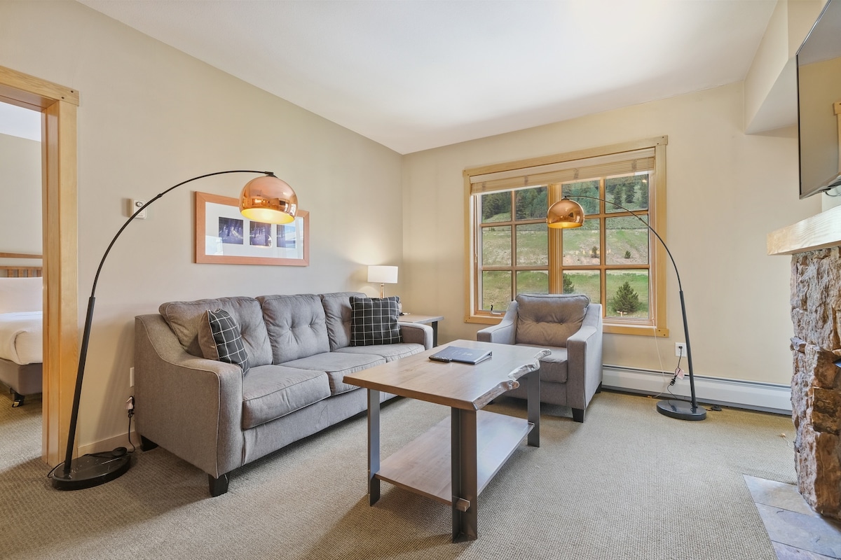 The living area features a grey sofa and a matching loveseat, complemented by two stylish floor lamps. A wooden coffee table sits in the center, with natural light flooding through a large window that offers views of the surrounding landscape.