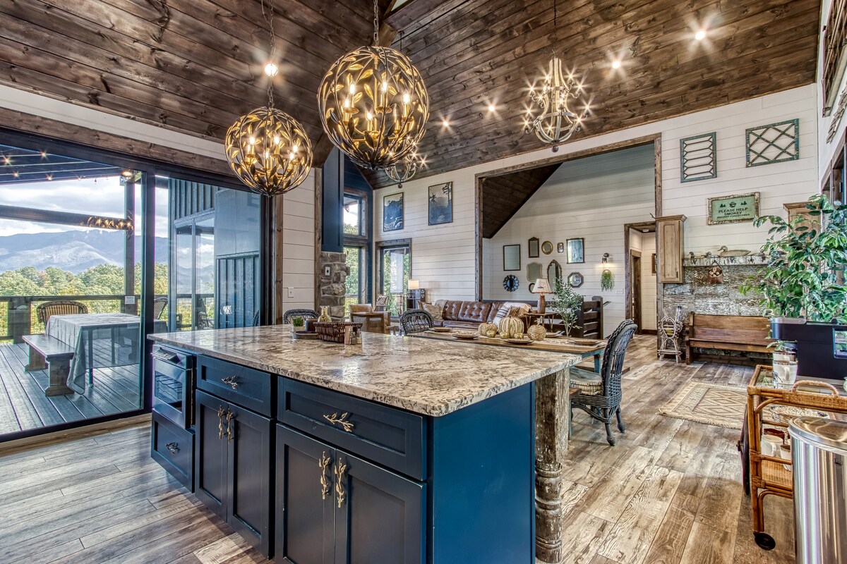 A stylish open-concept space features a large kitchen island with a speckled granite countertop. Unique pendant lights hang above, illuminating the area. Expansive glass doors provide views of the mountains and access to an outdoor deck. Various seating options are visible in the living area.