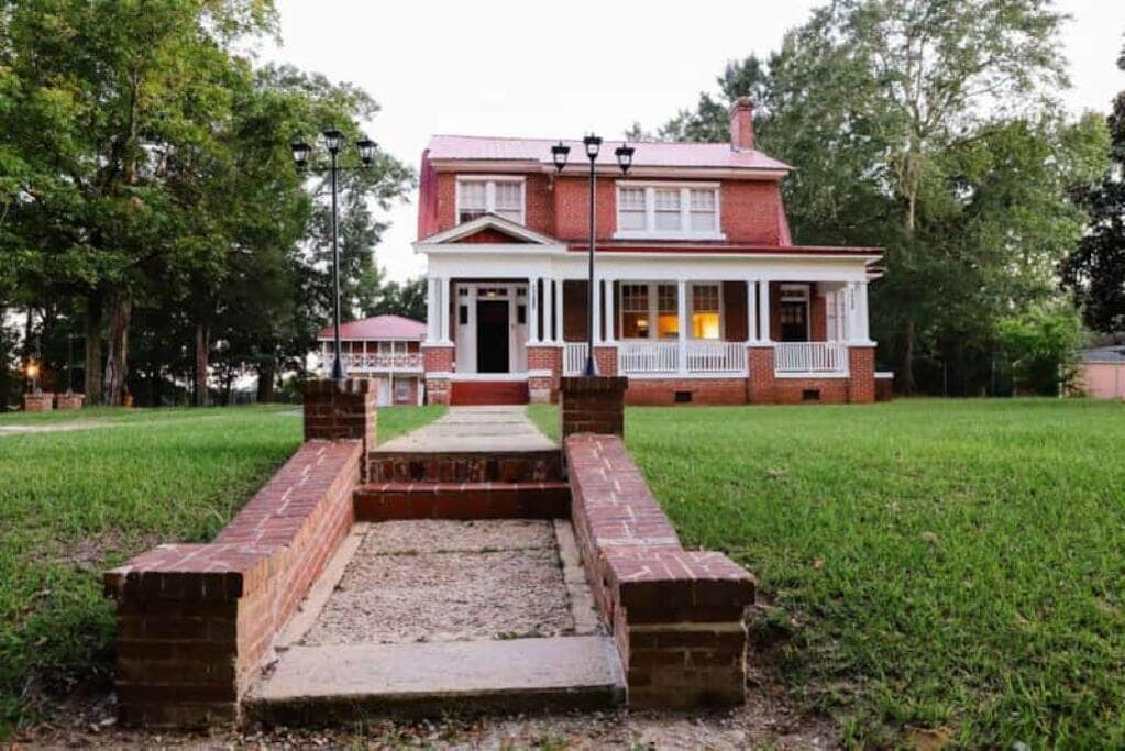 A historic estate is depicted, featuring a large red brick exterior and a welcoming front porch. Lush green grass surrounds the property, complemented by a well-maintained brick pathway leading to the entrance. Tall trees provide shade and enhance the serene atmosphere.