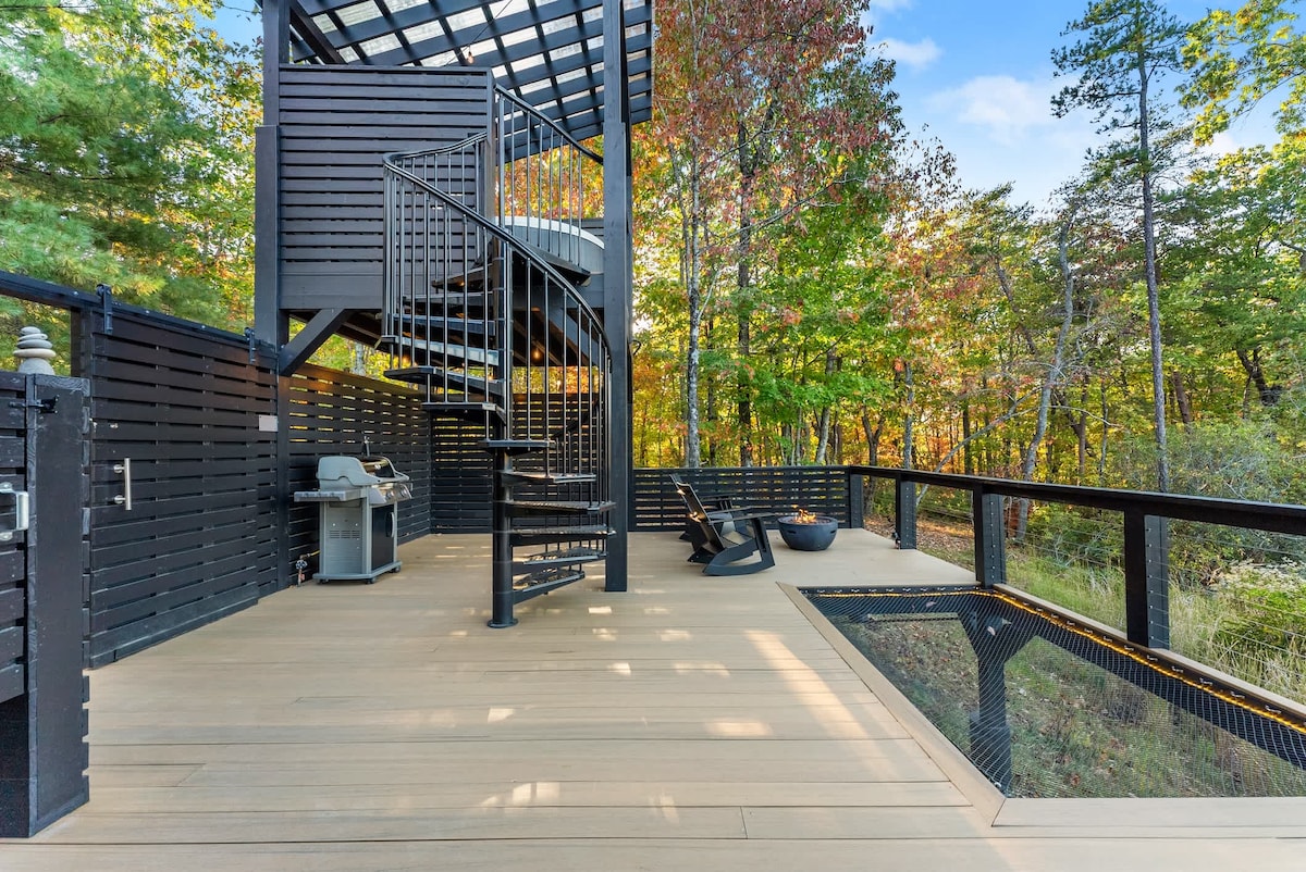 A spacious outdoor deck features a spiral staircase leading to an upper level. A gas grill is positioned against one wall, while a comfortable seating area and firepit invite relaxation amidst the surrounding trees and natural scenery.