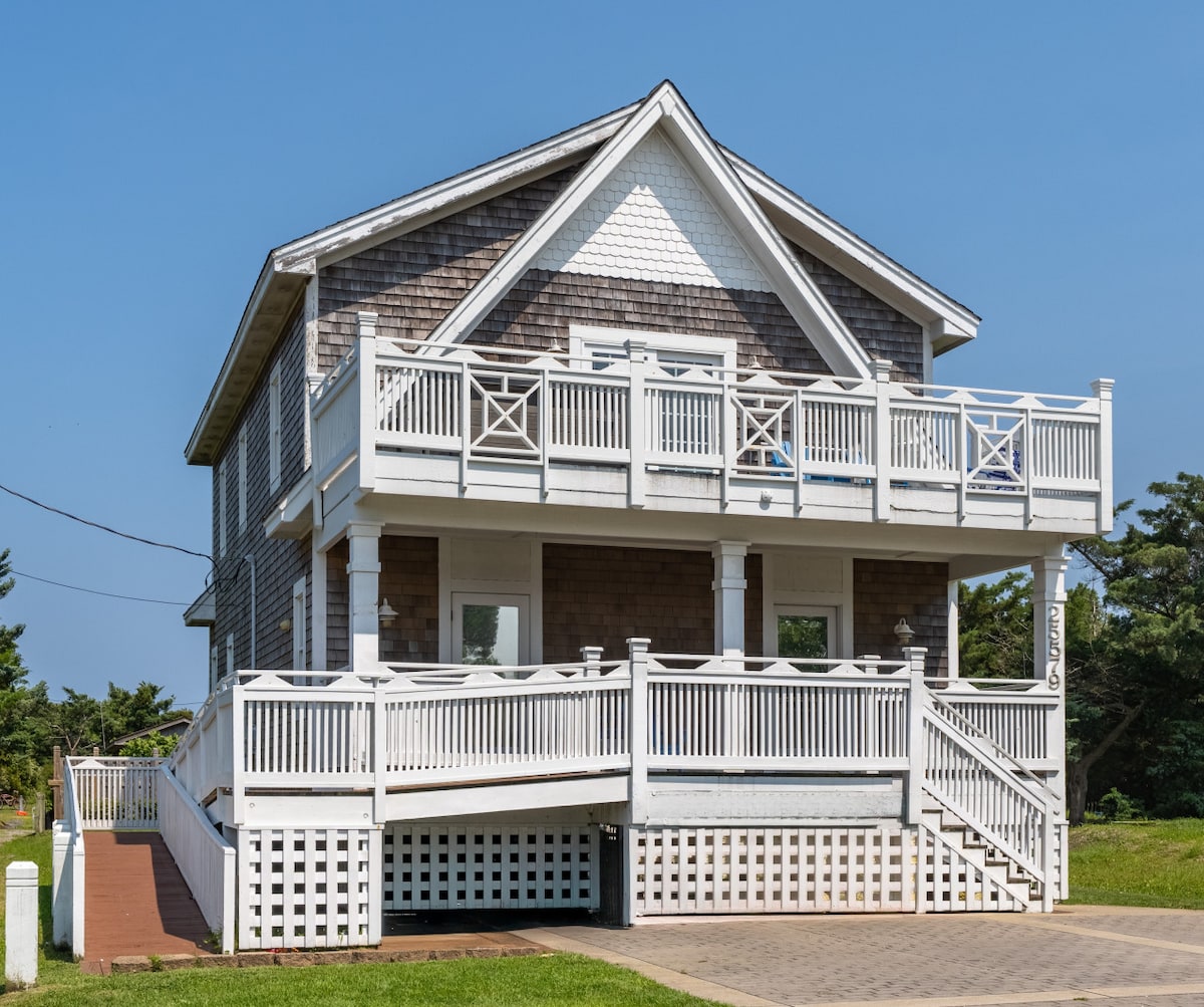 A charming beach house featuring a covered front porch with white railings and an inviting entrance. The two-story structure has a shingled exterior and multiple windows, allowing natural light to fill the interior. A well-maintained lawn surrounds the property, contributing to its welcoming presence.