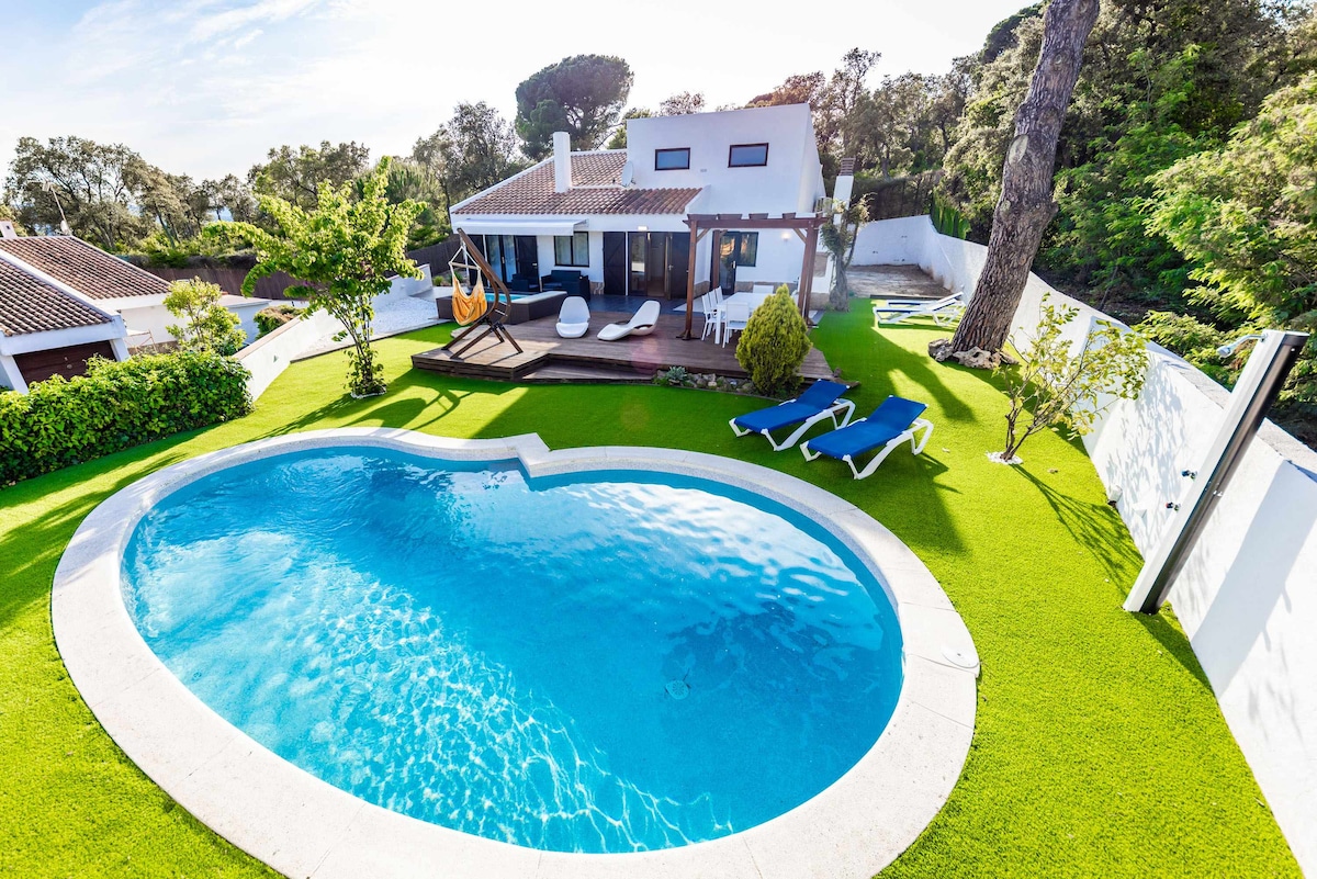 A heart-shaped swimming pool is surrounded by well-maintained green grass and lounge chairs. The outdoor space features a wooden deck with seating, providing a relaxed area to enjoy the views. The villa is partially visible in the background, complemented by trees.