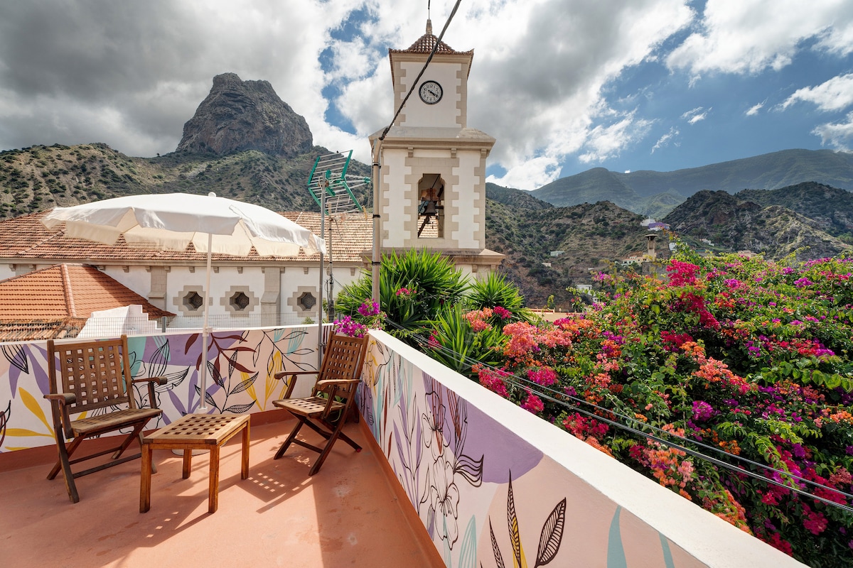 An open terrace features two wooden chairs positioned against a backdrop of vibrant, flowering plants. The mountains rise majestically in the distance under a partly cloudy sky, while a clock tower adds a charming element to the landscape.