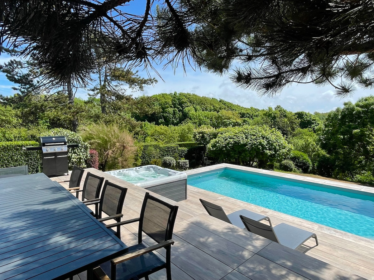 An outdoor space features a sleek heated swimming pool and a cozy jacuzzi, surrounded by lush greenery. A dining table with matching chairs offers a setting for meals, while sun loungers are positioned nearby, inviting relaxation. A barbecue grill is visible, enhancing the outdoor experience.