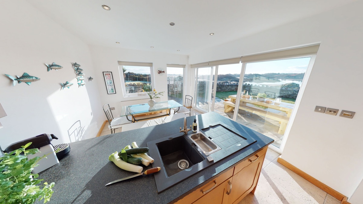 A modern kitchen features a large island with a sink, and a chopping knife resting next to a fresh vegetable. Ample natural light enters through expansive windows, revealing scenic views of the water and outdoor dining area with a wooden table.