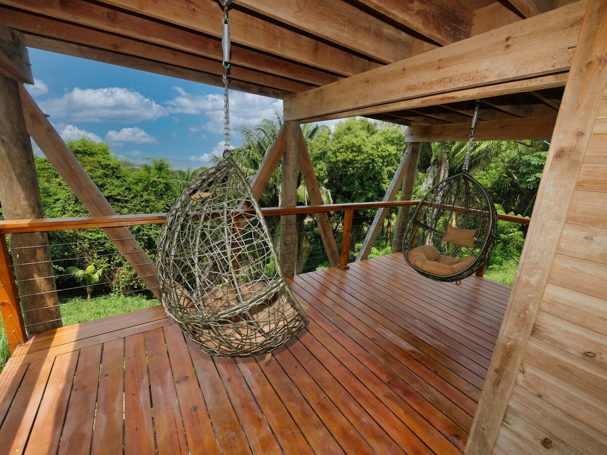 Two hanging egg chairs made of woven material are positioned on a wooden deck surrounded by lush greenery. The deck features wooden beams and flooring, providing a serene view of dense jungle trees under a bright blue sky.