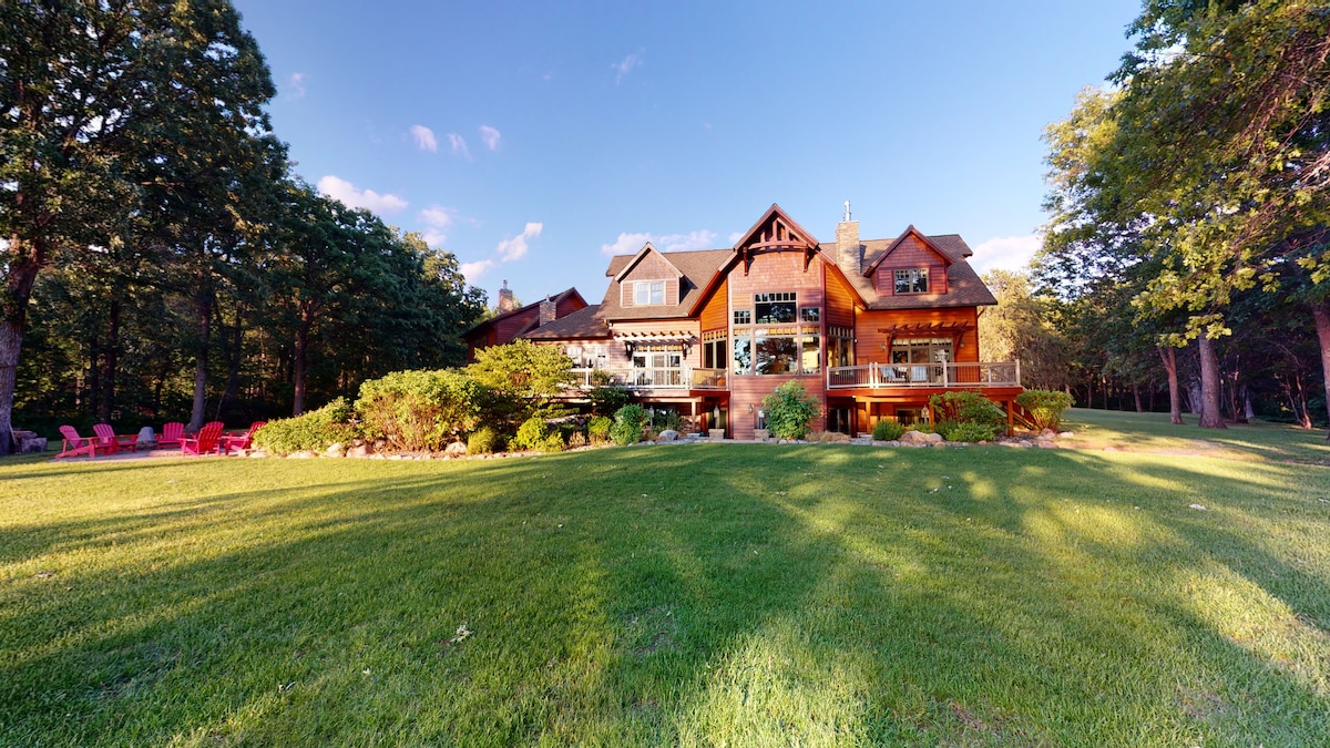 The estate is surrounded by lush greenery and gracefully landscaped areas, presenting a blend of natural beauty and inviting outdoor spaces. A spacious yard features vibrant grass and a cluster of seating options, while the multi-story wooden structure showcases large windows and a welcoming porch.