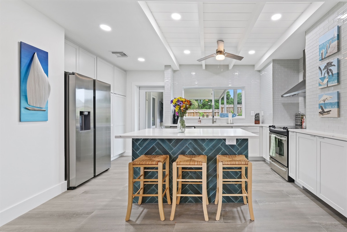 A modern kitchen features a large island with three wooden stools. Stainless steel appliances are positioned alongside white cabinetry. Natural light fills the space through a window above the sink, while nautical-themed artwork adorns the walls, adding a touch of coastal charm.