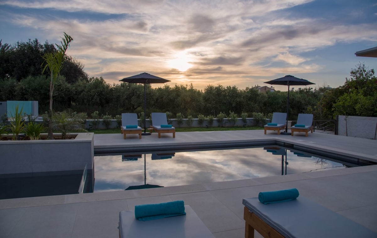 A serene pool area is captured at sunset, showcasing a still water surface reflecting the soft hues of the sky. Comfortable lounge chairs with blue towels are positioned nearby, while umbrellas provide shade. Lush greenery and planters surround the space, enhancing the tranquil ambiance.