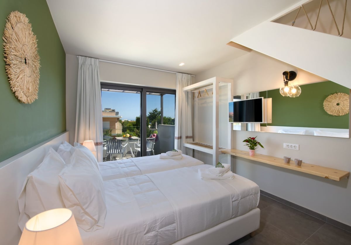 A modern loft features a cozy sleeping area with two spacious beds, complimentary white linens, and soft overhead lighting. Large glass doors open onto a private balcony, revealing greenery outside. A smart TV and a stylish mirror are positioned on the wall, enhancing the airy ambiance.