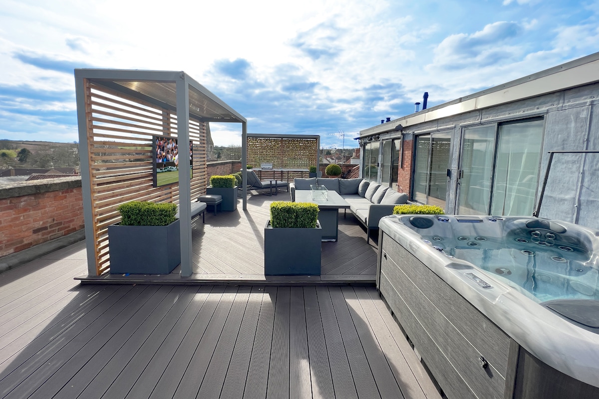 A spacious roof terrace features an outdoor lounge area with comfortable seating and a stylish dining table. A modern hot tub sits at one side, surrounded by concrete planters with greenery. An outdoor TV is mounted on a wooden structure, adding to the inviting atmosphere.