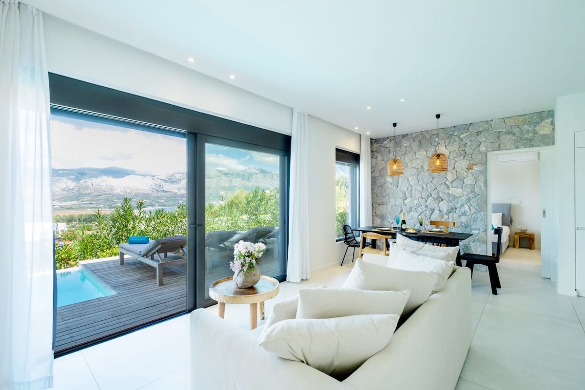 A light-filled living area features a comfortable sofa with plush cushions, large glass doors framing views of the pool and mountains. A dining table is set for meals, while natural stone walls provide a serene backdrop to the inviting space.
