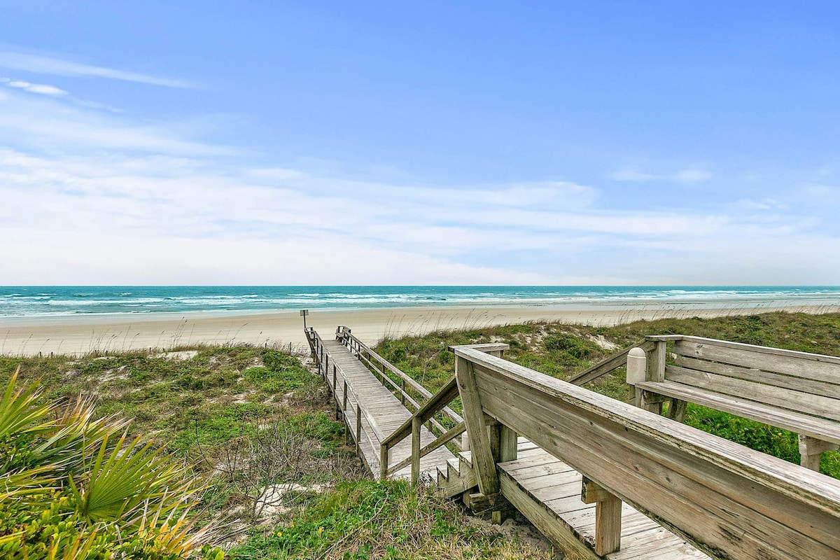 A wooden walkway leads from a grassy dune down to a sandy beach and turquoise ocean. Soft waves gently lap the shore under a clear blue sky, creating a serene coastal scene.