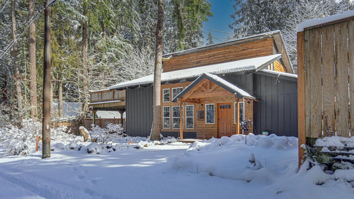 The exterior of Hearthstone Retreat is surrounded by a snowy landscape, showcasing a log and wood-paneled facade. A welcoming entrance with a large front porch is visible, flanked by large windows. Towering trees provide a natural backdrop, enhancing the serene setting.