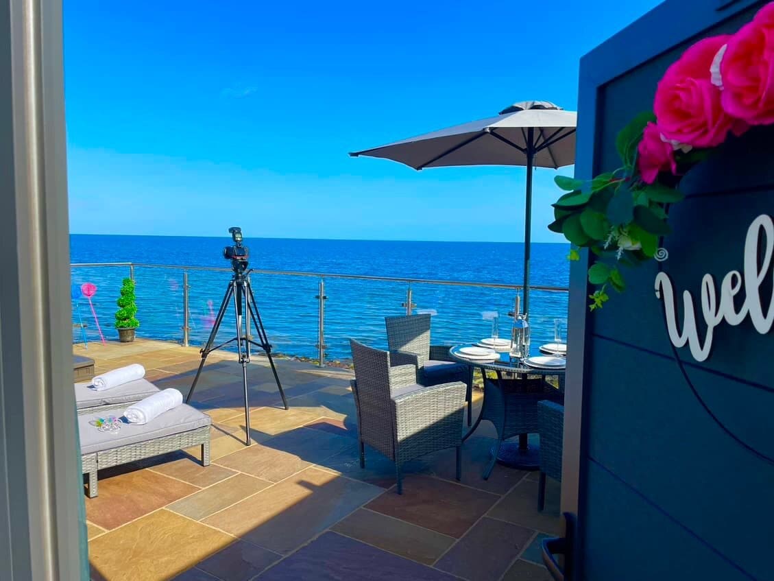 A private patio overlooks the sea, with seating arranged around a dining table. A parasol provides shade, and a telescope is positioned for viewing. The horizon features clear blue skies and gentle waters, enhancing the tranquil coastal setting.