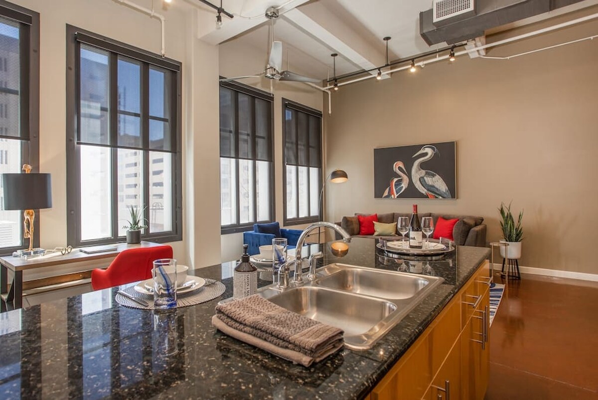 A modern kitchen area features a dark granite countertop with a double sink. Glassware and table settings are arranged for dining. Two stylish sofas in deep blue and red are positioned in the living space, accompanied by decorative plants and artwork on the wall.