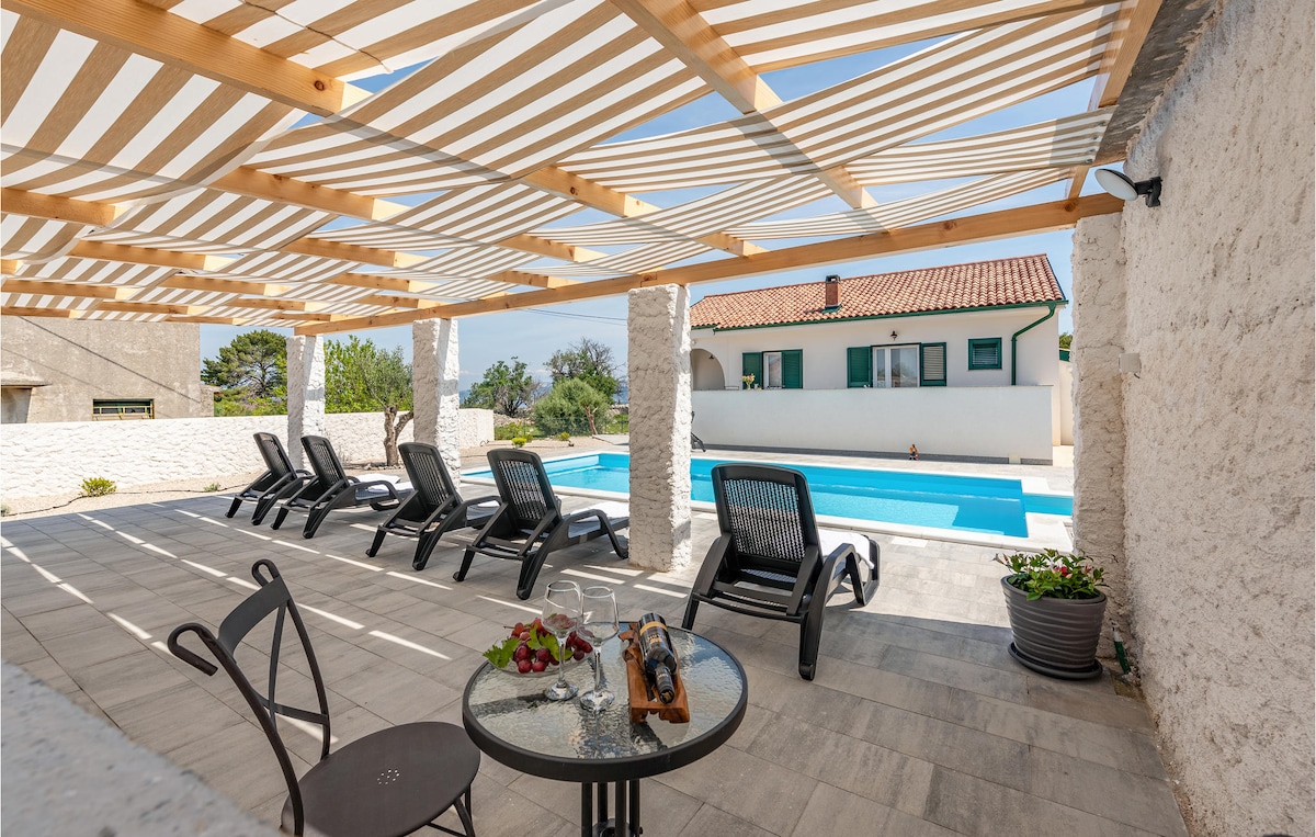 A shaded outdoor terrace features several loungers arranged around a private pool. A small table is set for refreshments, surrounded by a neutral-colored stone wall. The area is bright and spacious, ideal for relaxation, with trees visible in the background.