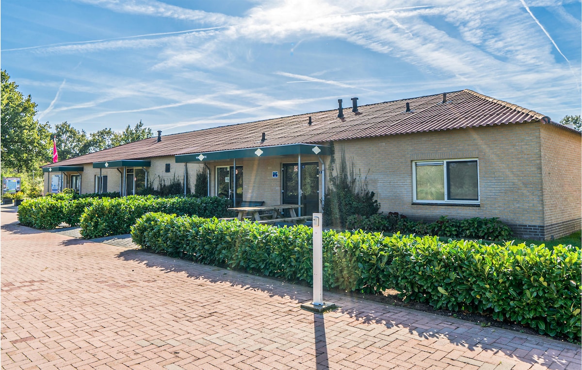 A single-story cottage is shown with a light-colored exterior and a sloping roof. Surrounding greenery includes well-maintained hedges and shrubs. The pathway is paved, leading to the entrance. Several large windows allow natural light into the space, enhancing the inviting appearance.