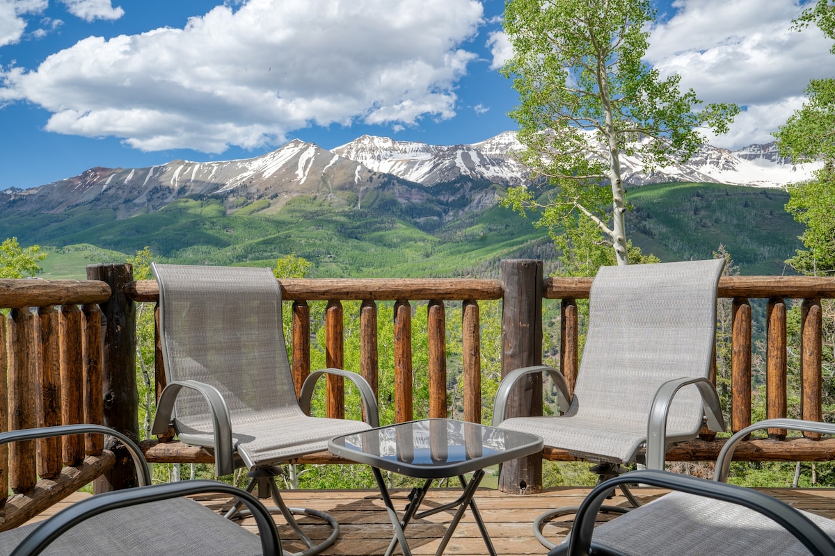 A wooden deck features two metal lounge chairs and a glass-top table, offering unobstructed views of snow-capped mountains and lush greenery. The sky is bright with fluffy white clouds, enhancing the scenic backdrop.