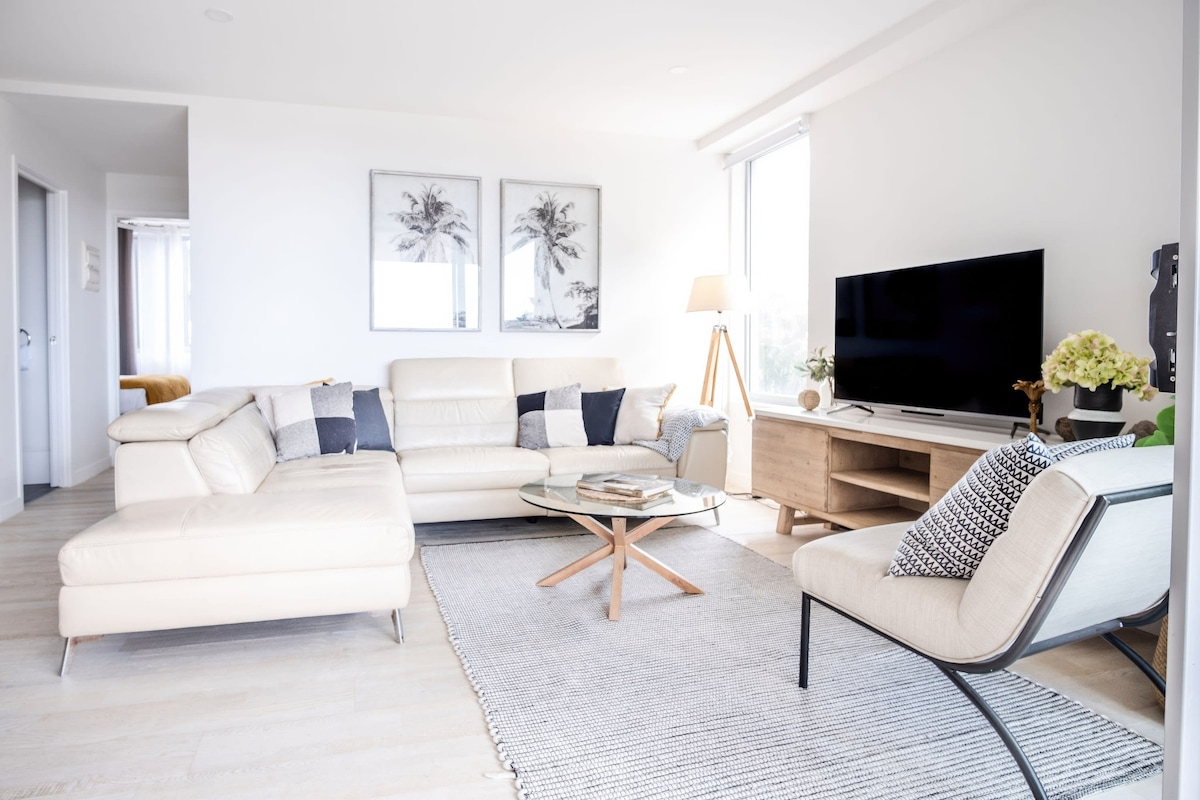 The living area features a spacious, light-colored sectional sofa complemented by an armchair. A glass coffee table is set on a woven rug, with a modern TV mounted on a wooden media console. Natural light is welcomed through large windows.