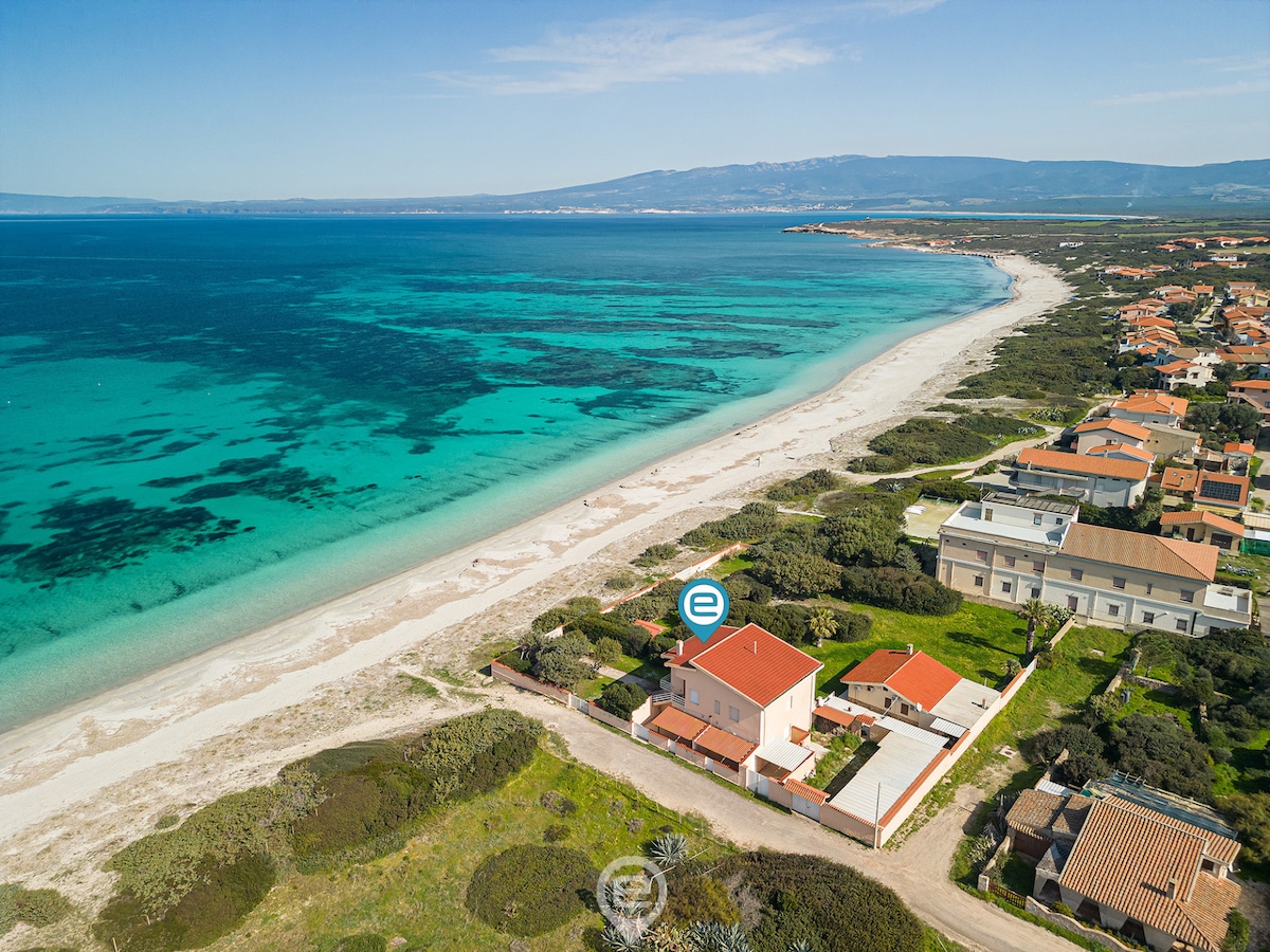 Aerial view showcasing House Minerva's proximity to Sa Rocca Tunda beach. The clear turquoise waters contrast with the fine sandy shore, while the spacious outdoor area of the house is seen alongside neighboring properties, emphasizing the tranquil coastal setting.