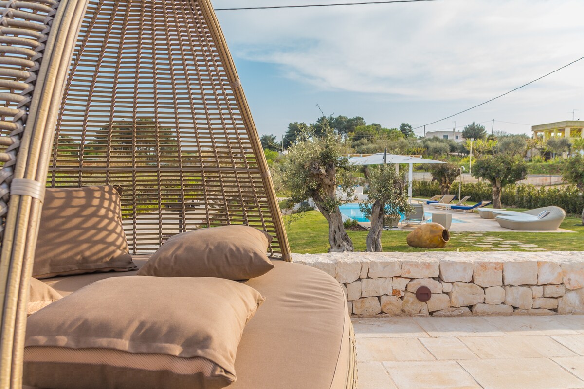 A comfortable seating area is framed by a wicker structure, featuring soft cushions. In the background, a lush garden is visible, with a heated pool and outdoor seating arrangements under a clear sky. Olive trees provide greenery to the serene outdoor space.
