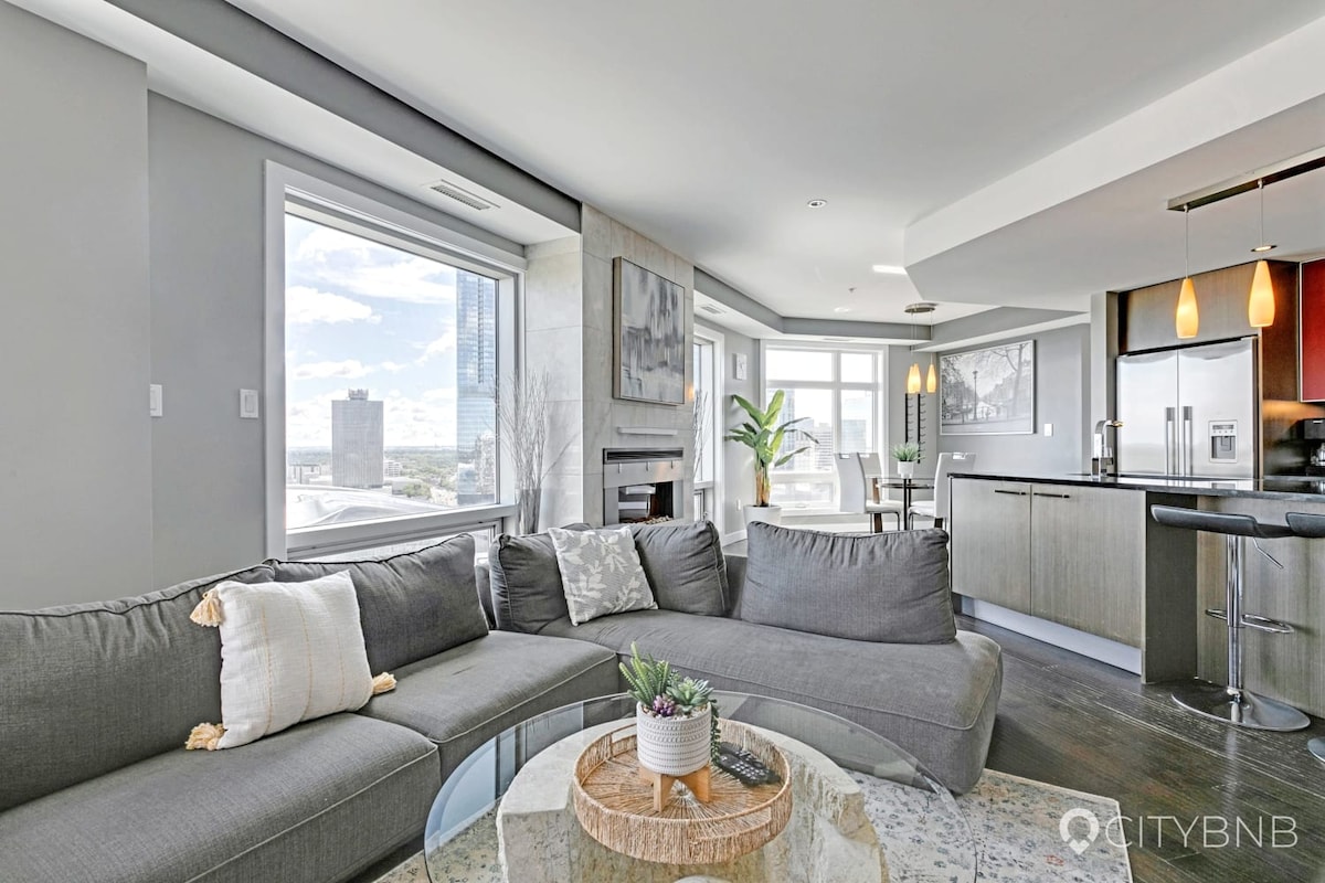 A spacious living area is highlighted by a contemporary sectional sofa and a round glass coffee table. Nearby, large windows provide ample natural light and a view of the city. The open kitchen features modern appliances and stylish pendant lighting.