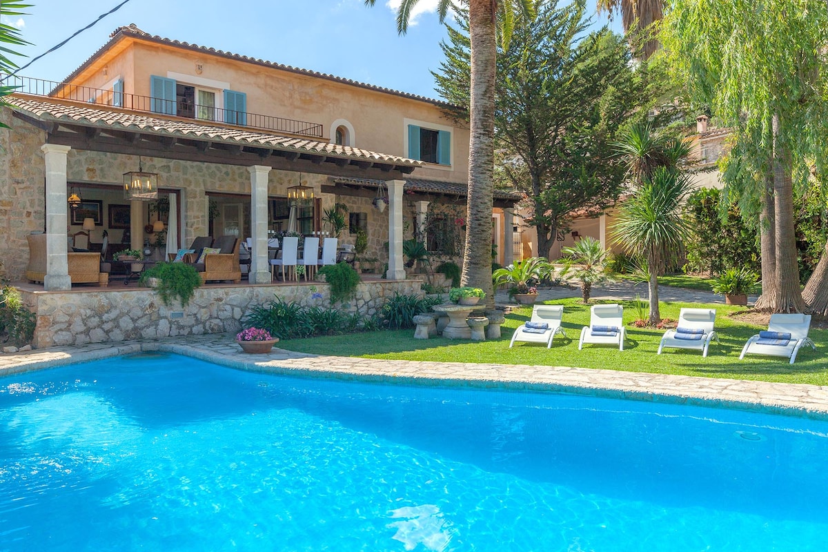 A private pool is framed by well-maintained gardens, featuring sun loungers arranged for relaxation. The villa's covered veranda, furnished with seating, provides a scenic view of the property and the vibrant greenery surrounding it.