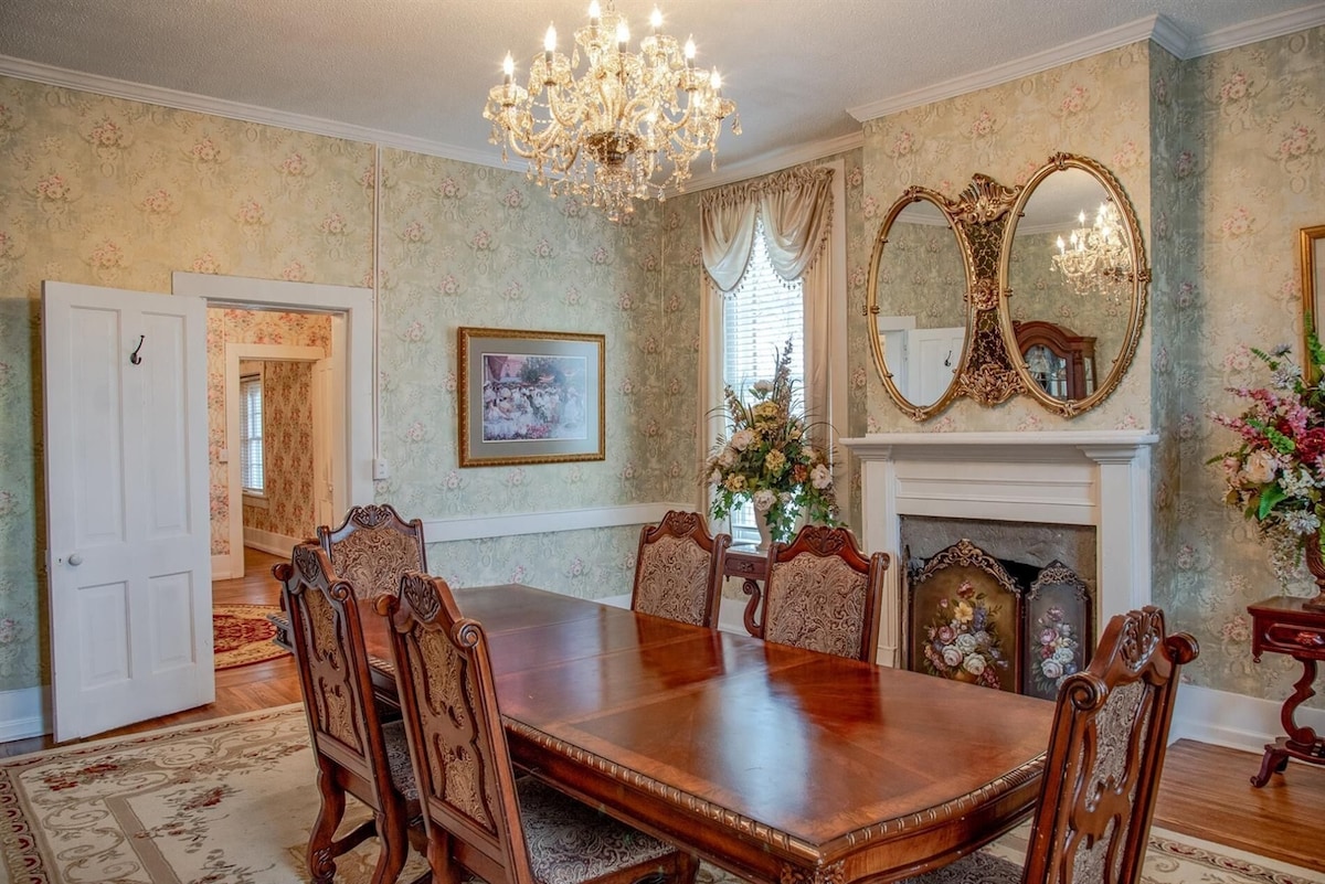 A formal dining room features a large wooden table surrounded by intricately designed chairs. Elegant wall coverings in soft florals are complemented by a crystal chandelier above. Mirrors and a decorative fireplace add to the room's classic charm, while natural light enters through a window draped with curtains.