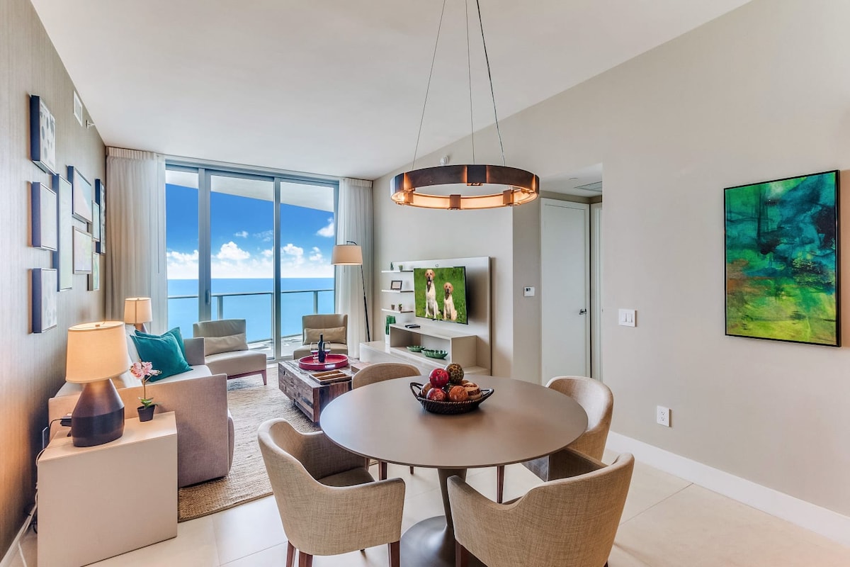 A living and dining area features a round table surrounded by four upholstered chairs. A soft sofa and a coffee table are positioned nearby. Floor-to-ceiling glass doors offer a view of the ocean, while a vibrant artwork piece adds a pop of color to the neutral-toned walls.