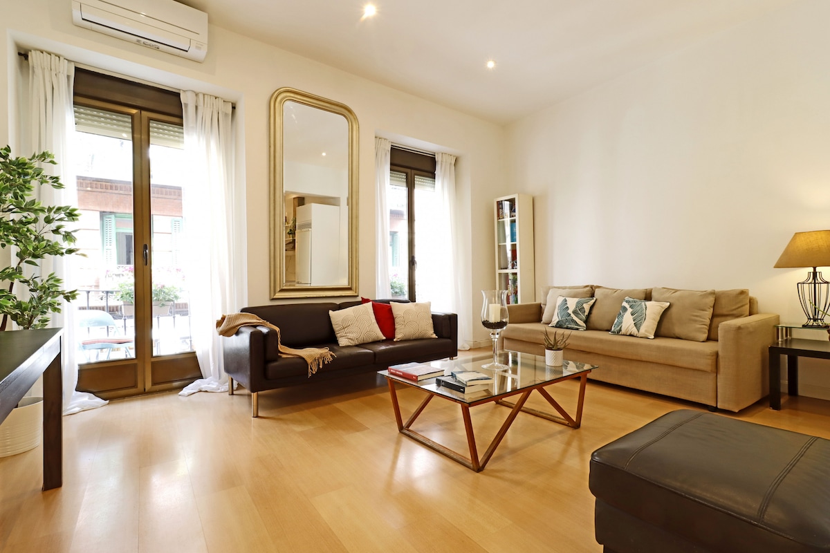 A bright living area is showcased with light-colored walls and wooden flooring. A large mirror is positioned above a coffee table, surrounded by a dark sofa and a beige couch. Natural light filters through the balcony door, illuminating decorative cushions and a lamp.