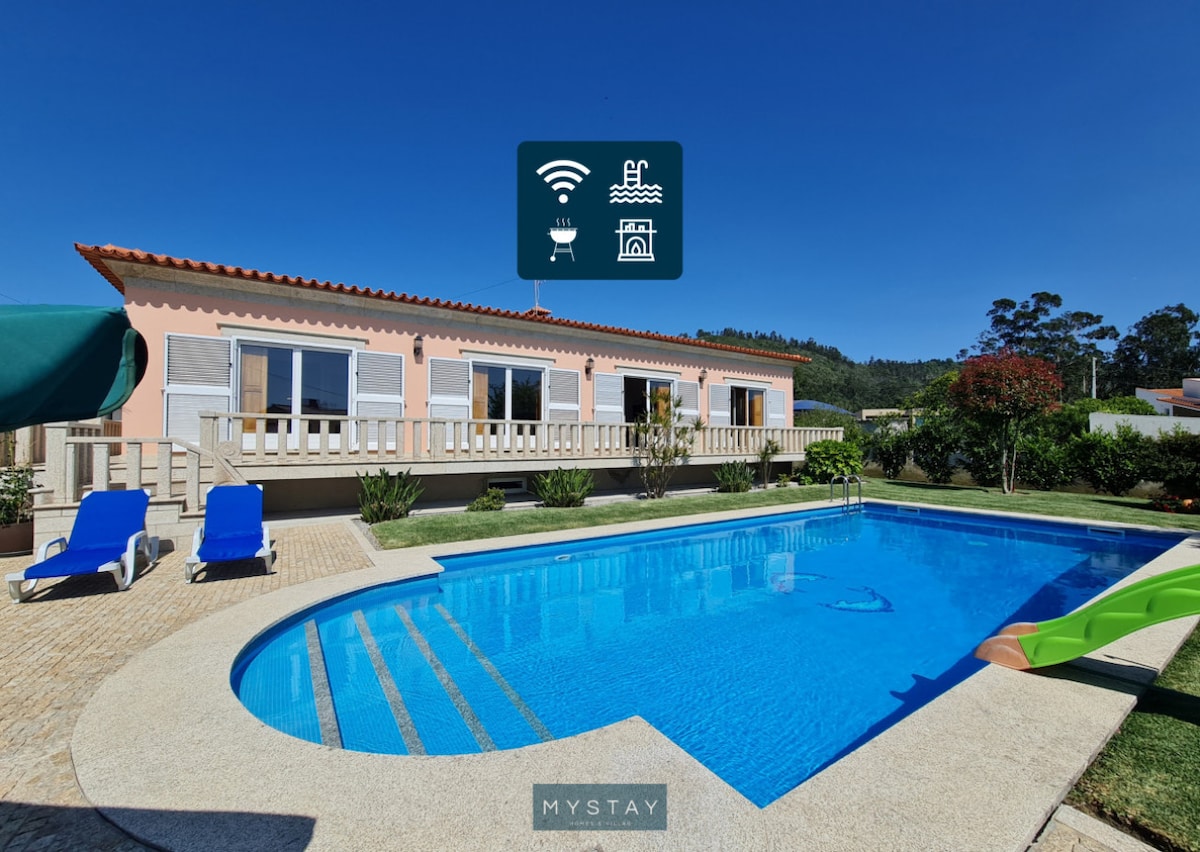 A luxurious villa features a private swimming pool surrounded by a well-maintained garden. Sun loungers are positioned near the pool, while an outdoor dining area is visible on the patio. The spacious façade of the house offers large windows that enhance natural light.
