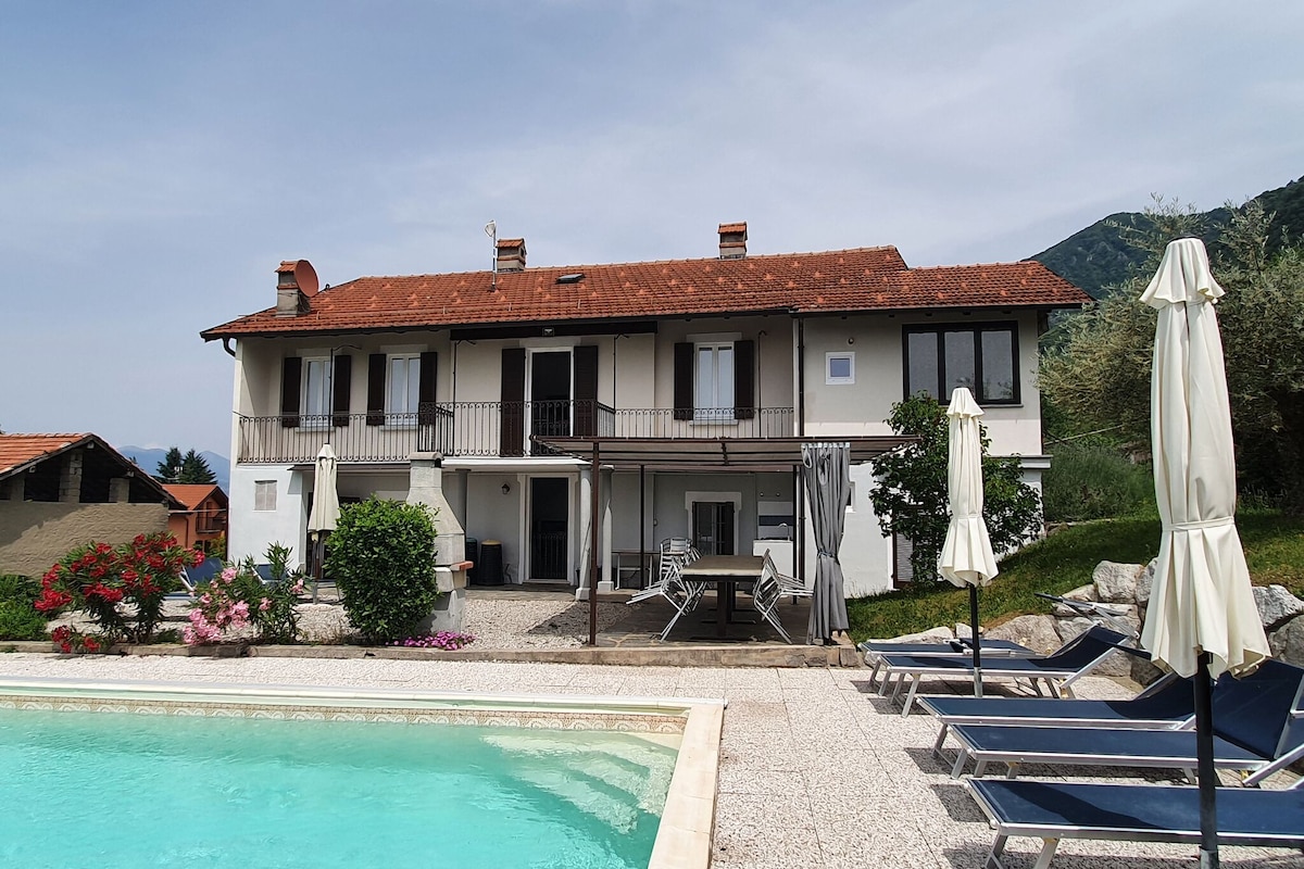 A two-story holiday home is displayed, featuring a spacious outdoor area with a private swimming pool. Lounge chairs are arranged beside the pool, and shaded umbrellas provide comfort. The building has a mix of windows and balconies, surrounded by greenery and colorful flowers.
