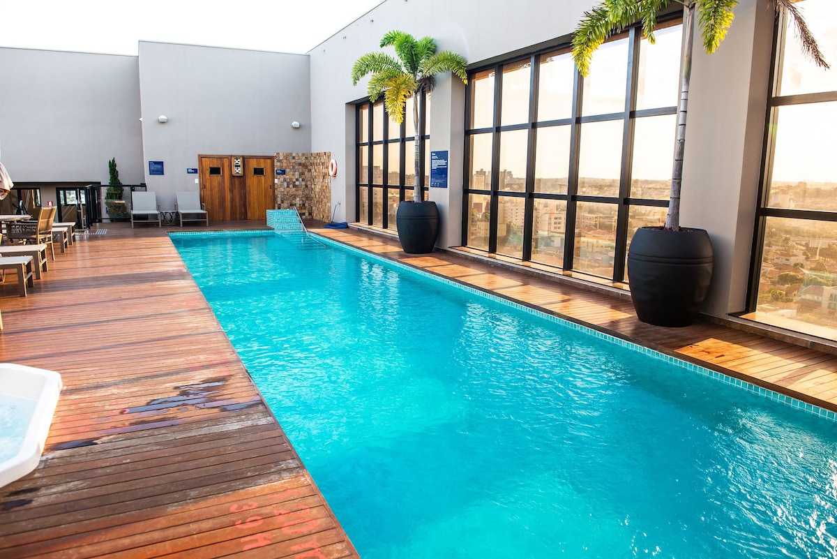 A rooftop pool area features a clear, blue swimming pool surrounded by wooden decking. Large windows allow for ample natural light, and two potted palm trees add a touch of greenery. The tranquil setting is designed for relaxation.