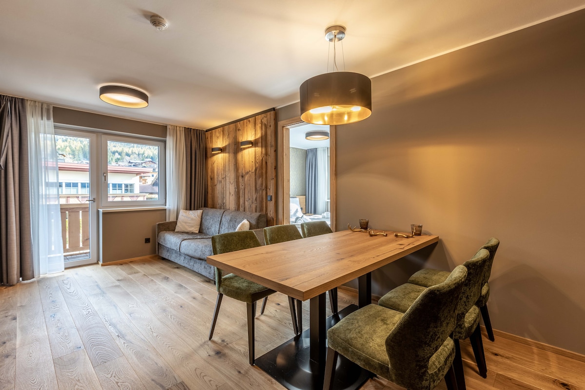 A dining area features a wooden table surrounded by four green chairs. A gray sofa sits against a wall with wood paneling. Natural light enters through the large balcony door, illuminating the space. Ceiling lights provide additional warm lighting.