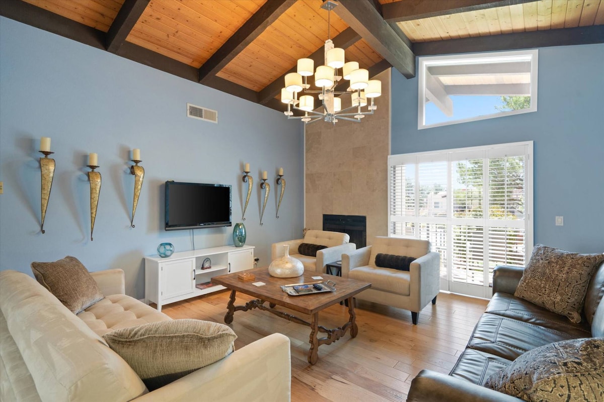The living room features a high, exposed beam ceiling with a warm wooden finish. Soft seating is arranged around a wooden coffee table, with decorative elements on the walls and a modern television mounted above a white console. Natural light enters through large windows.