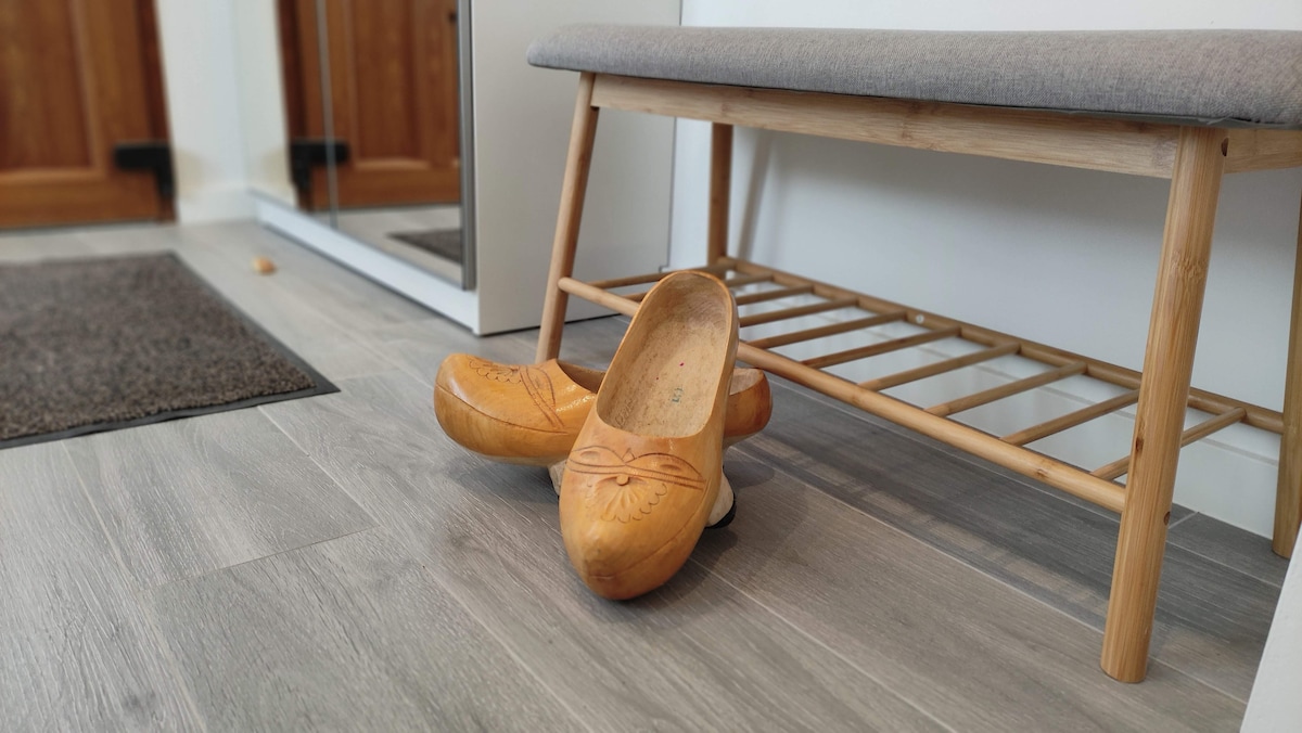 A wooden bench with a gray cushion is positioned against a wall, complemented by a shoe rack underneath. Traditional wooden clogs rest on the floor nearby, with light-colored flooring enhancing the room's clean and organized appearance.