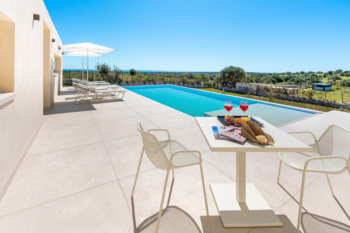 An outdoor terrace features a table set for two with refreshments, surrounded by two chairs. A spacious infinity saltwater pool is positioned in the background, bordered by a scenic landscape of greenery and distant hills, and shaded by two umbrellas.