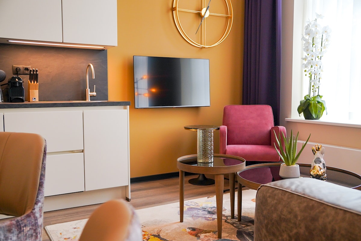 An inviting living area is highlighted by a warm orange wall. A modern kitchen unit with a coffee machine is positioned nearby. A plush pink armchair and a coffee table are featured in front of a flat-screen TV, complemented by decorative plants on the tables.