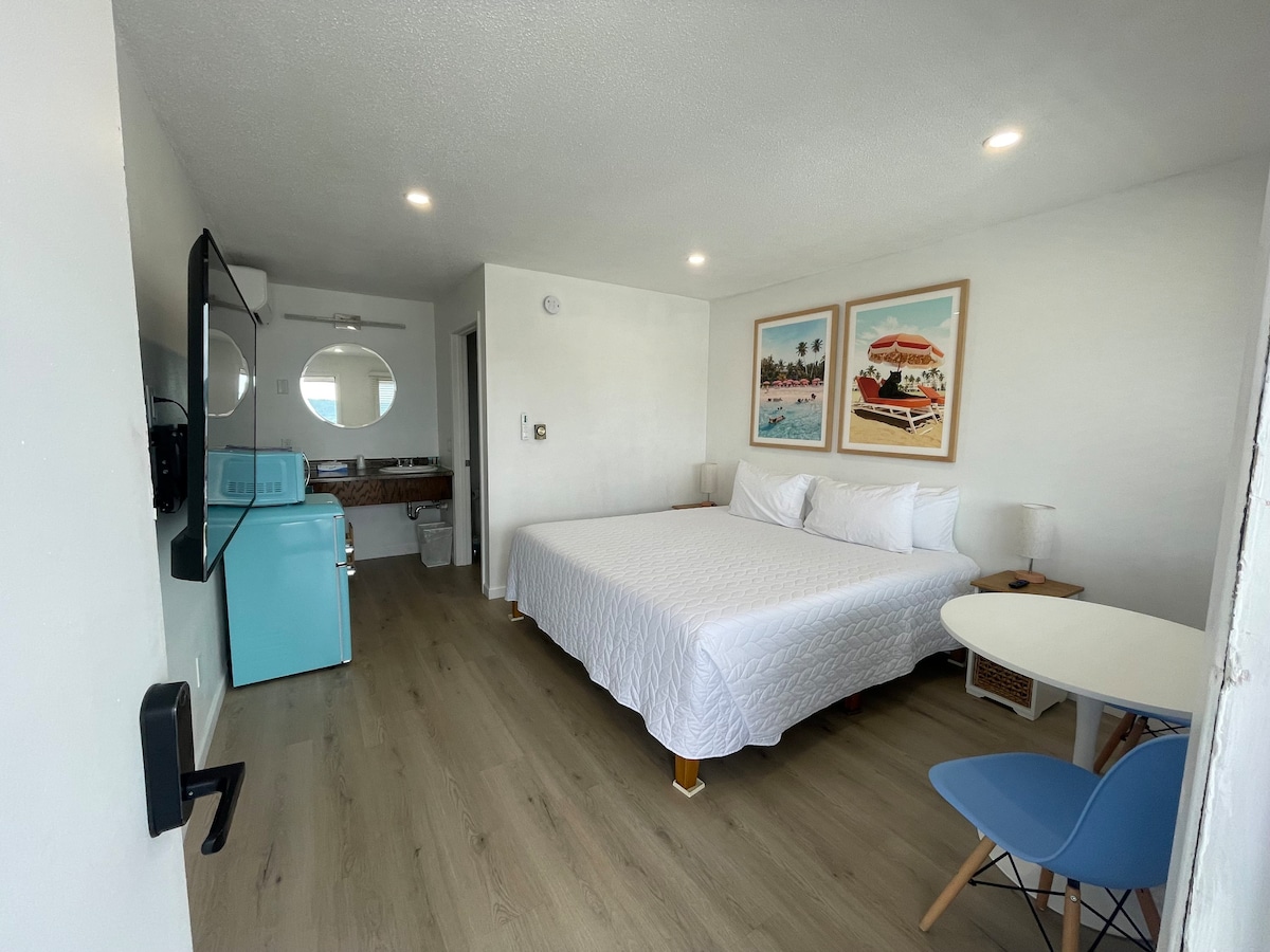 A well-lit room features a spacious bed with crisp white linens. Two vibrant paintings adorn the wall above a small nightstand, and a blue retro mini-fridge adds a pop of color. A seating area with a blue chair is positioned near the window, enhancing the inviting space.