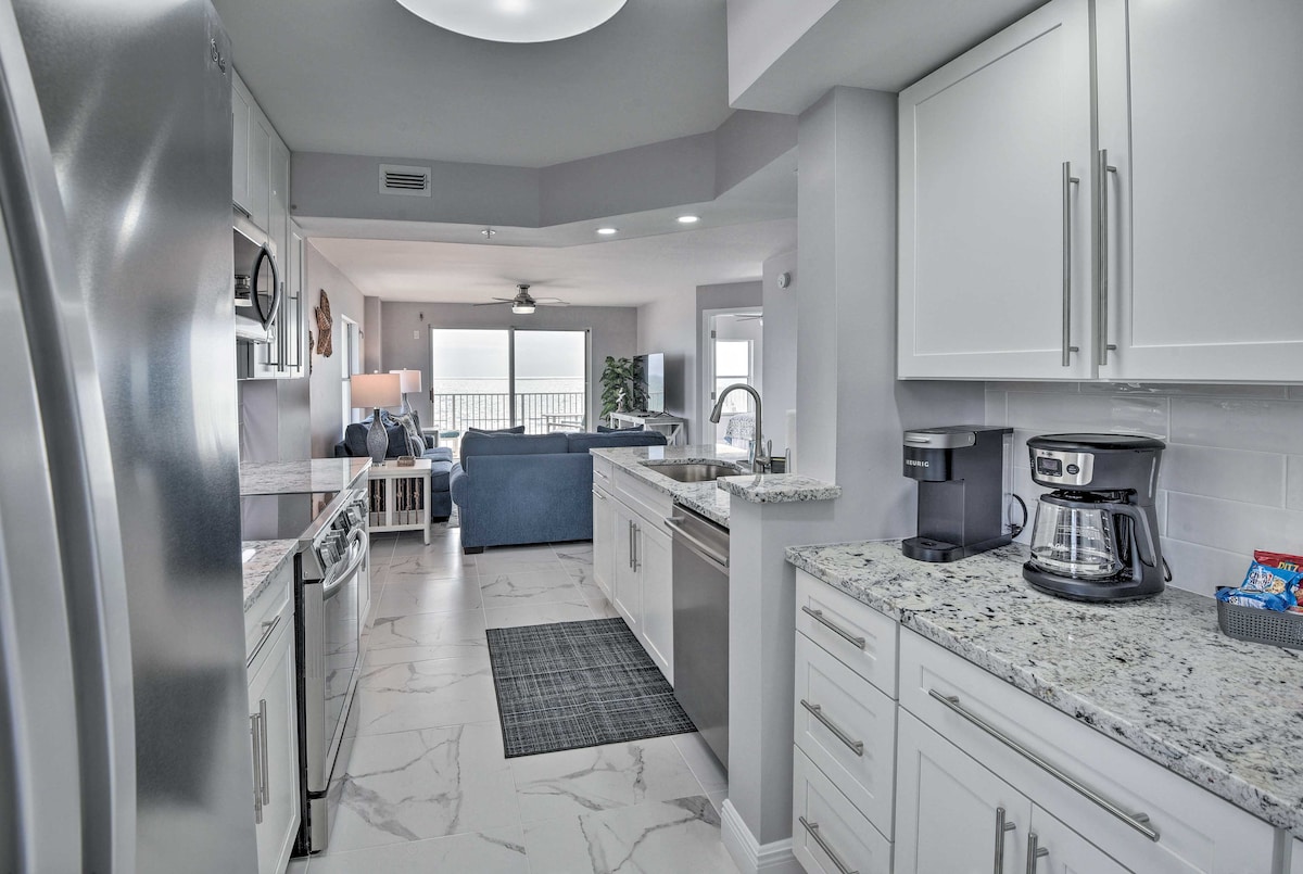 A bright, modern kitchen is showcased with stainless steel appliances and granite countertops. The space includes a large sink, ample storage, and a coffee maker. A cozy dining area is visible, with a view of the ocean through nearby windows that allow natural light to fill the room.